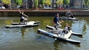 Drie mensen op een Schiller Waterfiets in een gracht van Delft, met op één fiets een passagier. Foto: Foto: BootjeVarenDelft