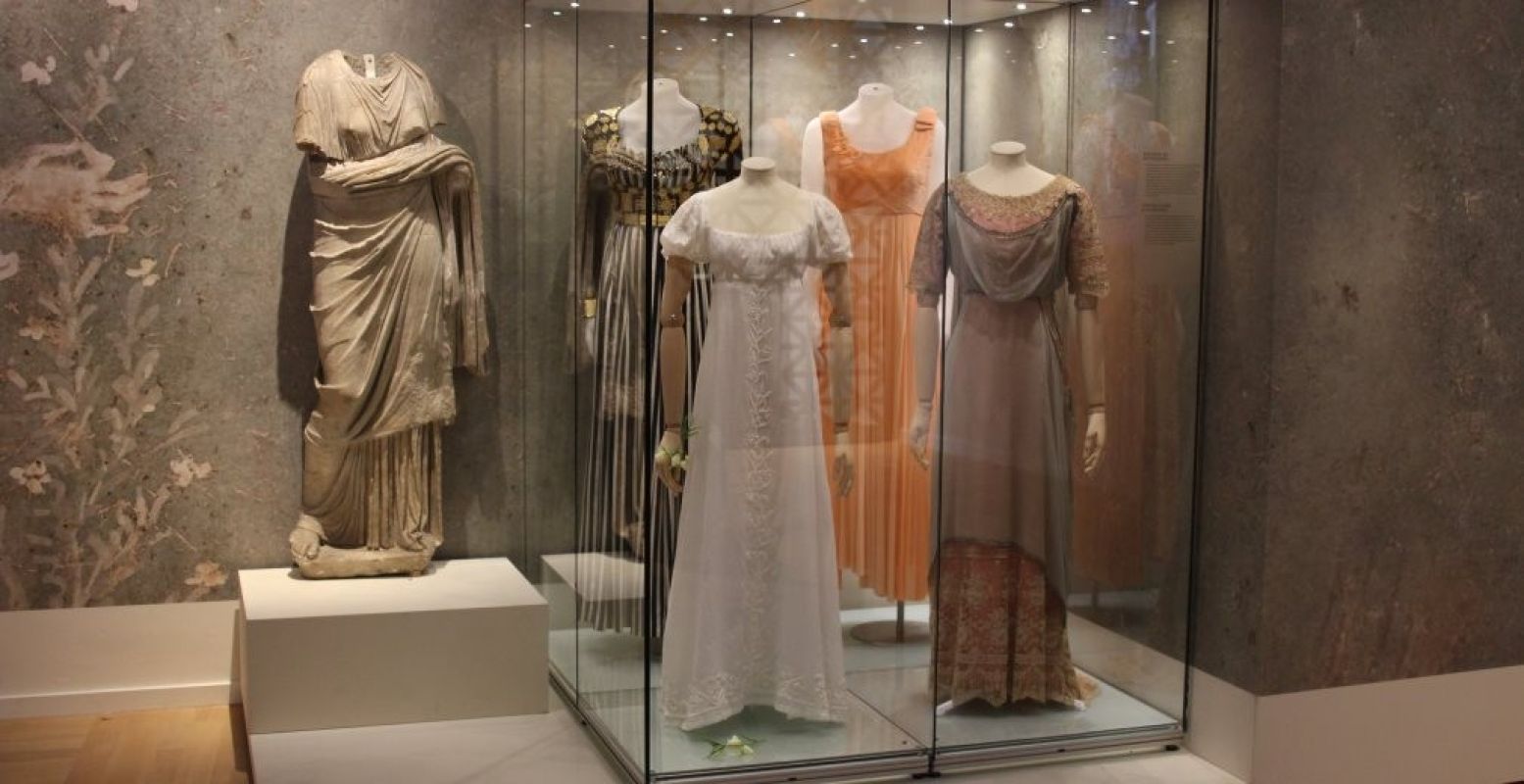 Prachtige jurken in Empirestijl; geïnspireerd op de damesmode van de Romeinen zoals het standbeeld naast de vitrine laat zien. Foto: Redactie DagjeWeg.NL
